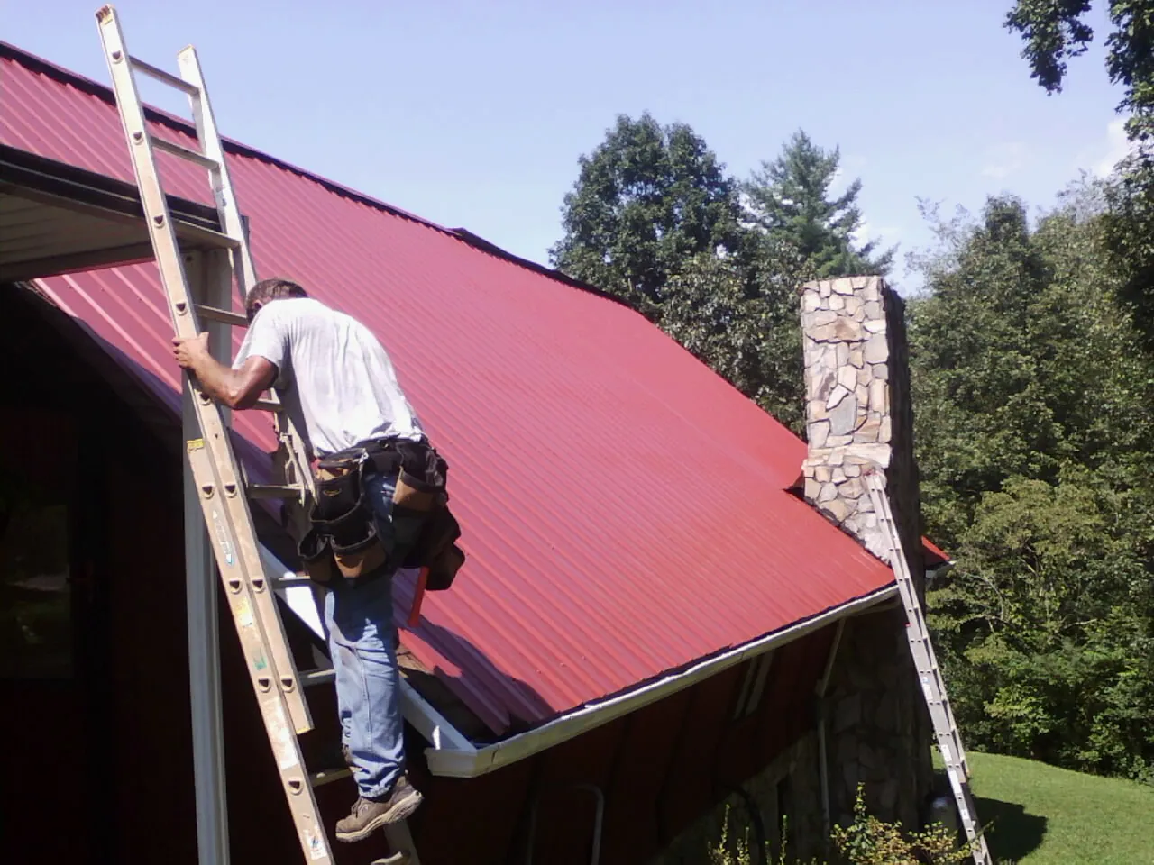 Roofer on a ladder beside a red metal roof and stone chimney — Soffit & Fascia Repair by Roofing Brothers in Hardyville